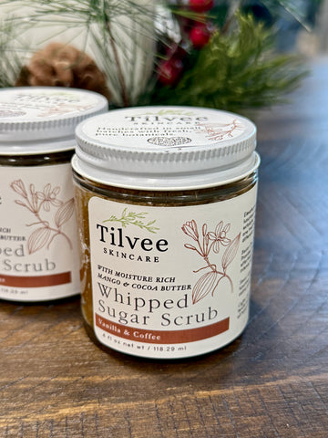 Two jars of Tilvee Skincare Whipped Sugar Scrub on a wooden surface with a blurred background.